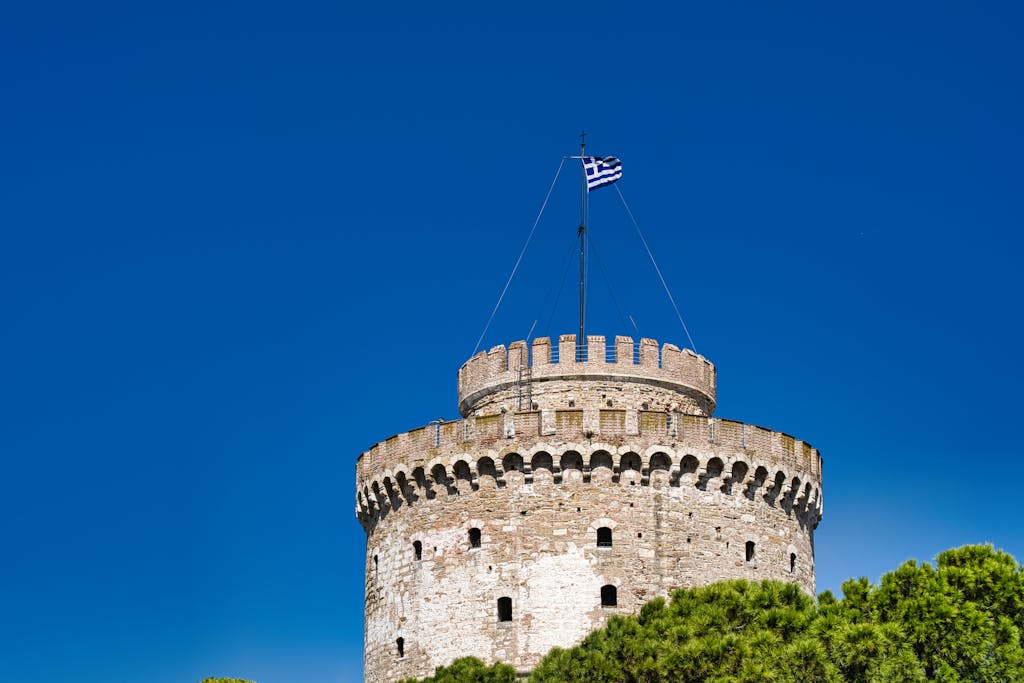 Majestic view of the historic White Tower of Thessaloniki against a bright blue sky with a Greek flag.