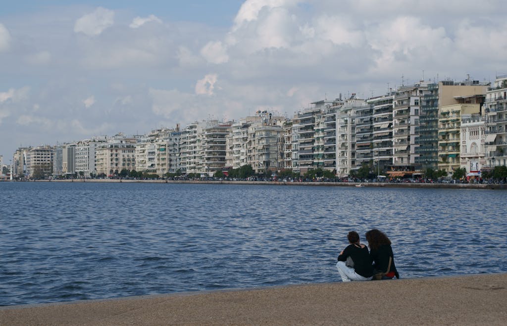 Two women sitting by the sea with a view of urban buildings in Thessaloniki, Greece.