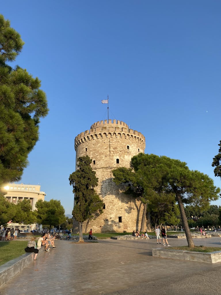 View of the iconic White Tower in Thessaloniki, Greece, under clear blue skies with surrounding trees.